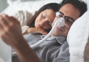 Woman sleeping next to her husband while he reads in bed while wearing his CPAP nose mask