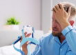 Man sitting on the bed with his hand on his head appearing frustrated and holding a blue CPAP mask.