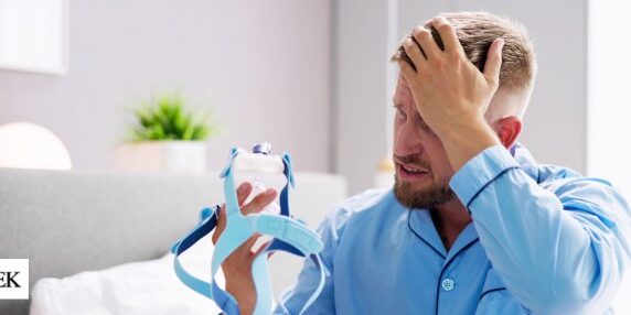 Doubek-CPAP Man sitting on the bed with his hand on his head appearing frustrated and holding a blue CPAP mask.
