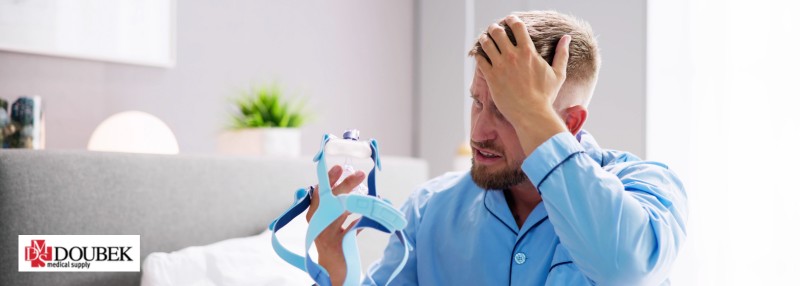 Man sitting on the bed with his hand on his head appearing frustrated and holding a blue CPAP mask.
